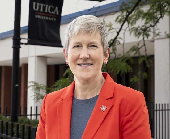 Provost Stephanie Nesbitt, in orange blazer, smiles in front of DePerno Hall on the Utica campus.