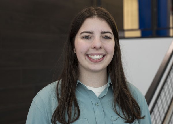 Sydney Kinsella, with long dark hair and wearing a blueish-green button-down top, looks at the camera and smiles.