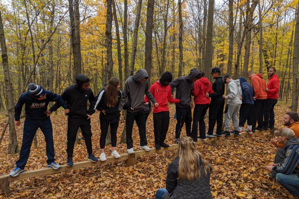 Students in the woods stand on a log holding hands.