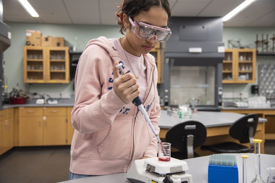 Yamilet Taveras ’27, a chemistry major wearing a pink hoodie and safety goggles, pours a substance into a beaker in the chemistry lab.